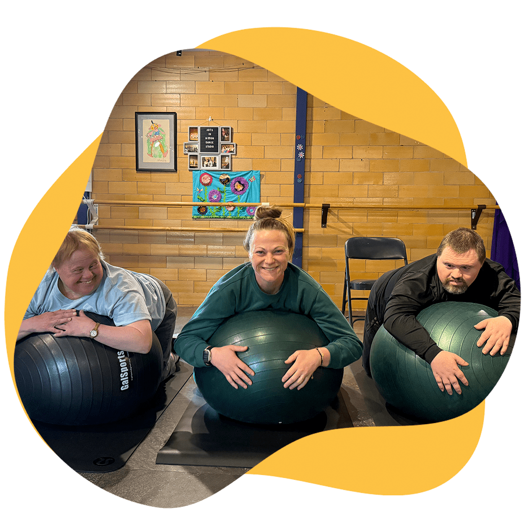 Two students with yoga instructor resting on large rubber exercise balls while smiling for a photo in the studio.